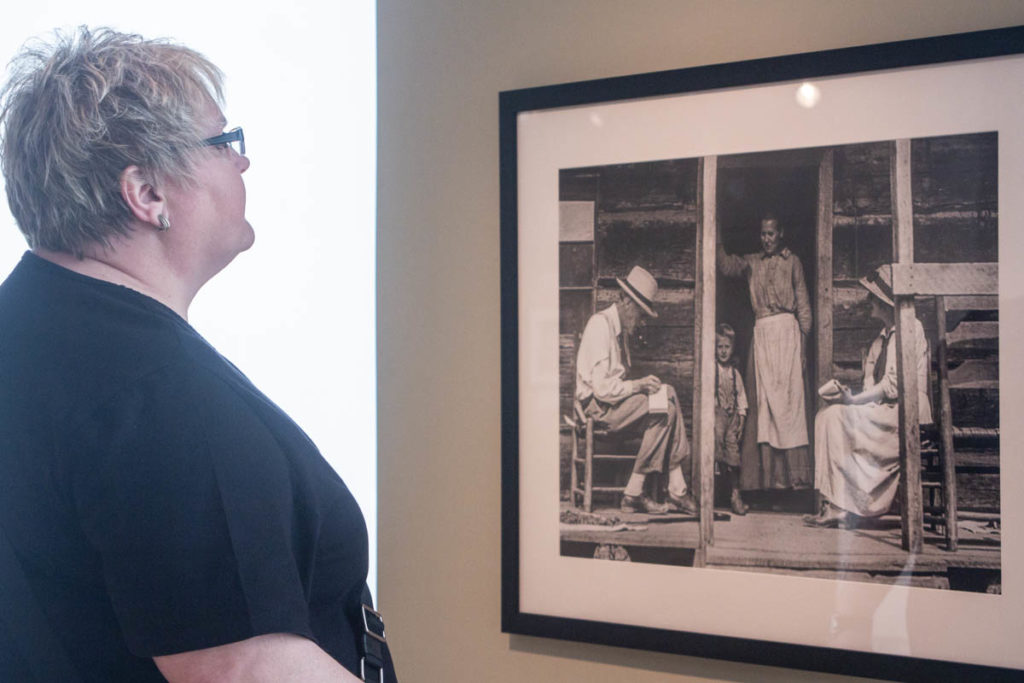 A visitor looks at a photograph of Cecil Sharp and his assistant recording the words/lyrics of an Appalachian woman on her porch. A small child stands in the doorway with her.