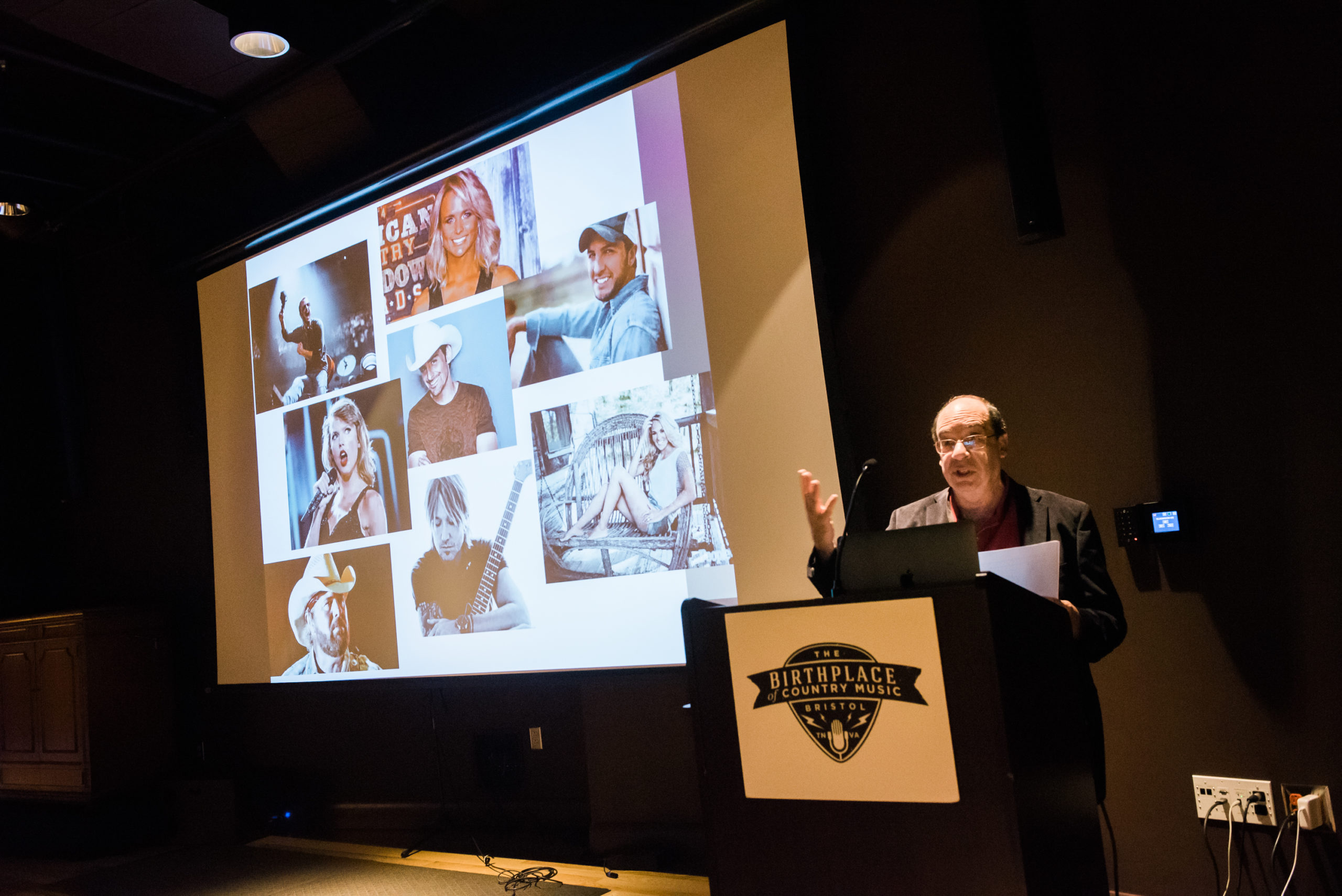 Barry Mazor stands at a podium in a darkened theater with a screen behind him that shows several pictures of different country music musicians and stars.