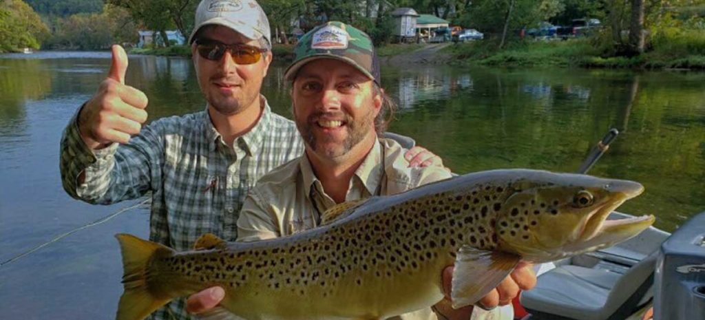 2 fishermen showing off the catch of the day for the camera. One man is giving a thumbs up, the other is holding a nice sized fish.
