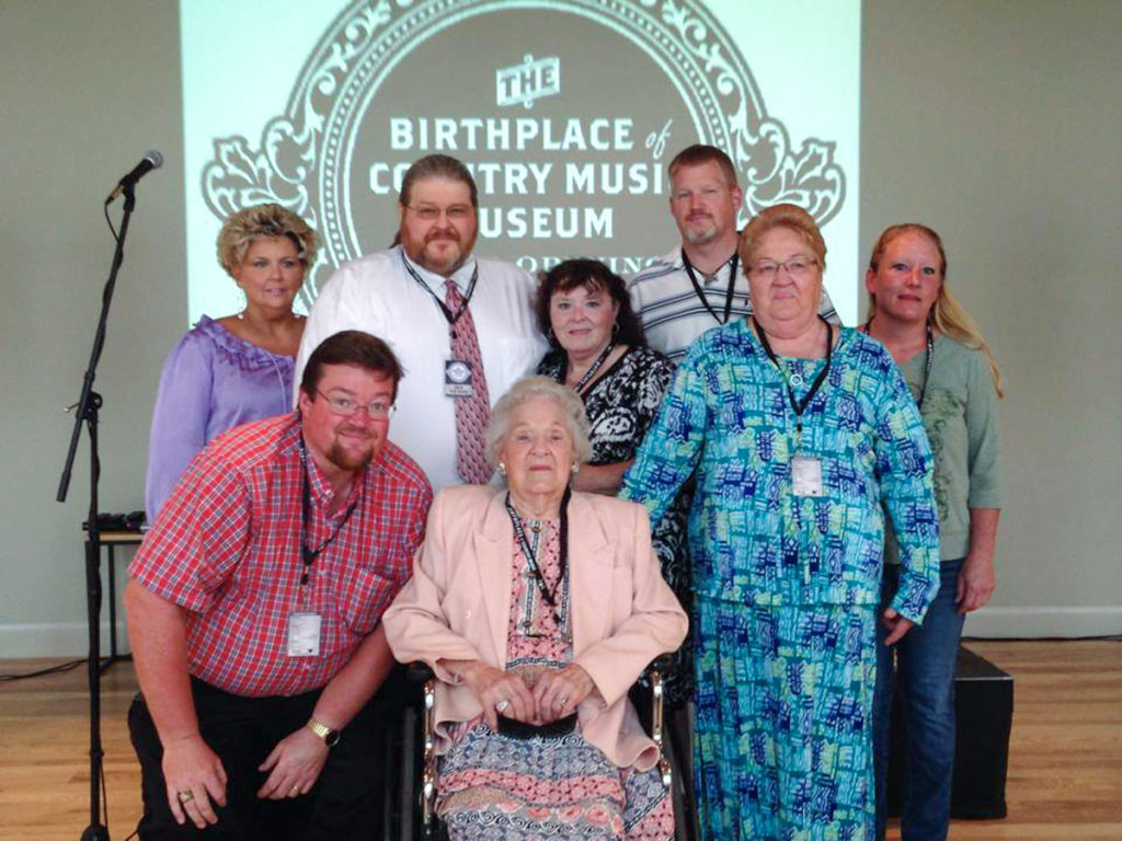 Georgia Warren sits central, surrounded by her daughter Nancy and several other family members. Behind them is the Grand Opening Birthplace of Country Music Museum logo.