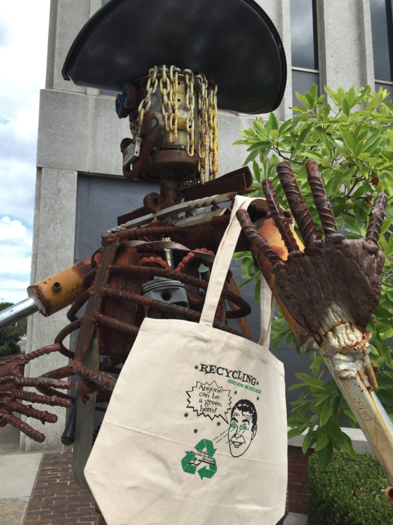 The metal statue of Dale, always on display at festival, wearing a Green Team canvas tote bag with one of the t-shirt designs about being a green hero.