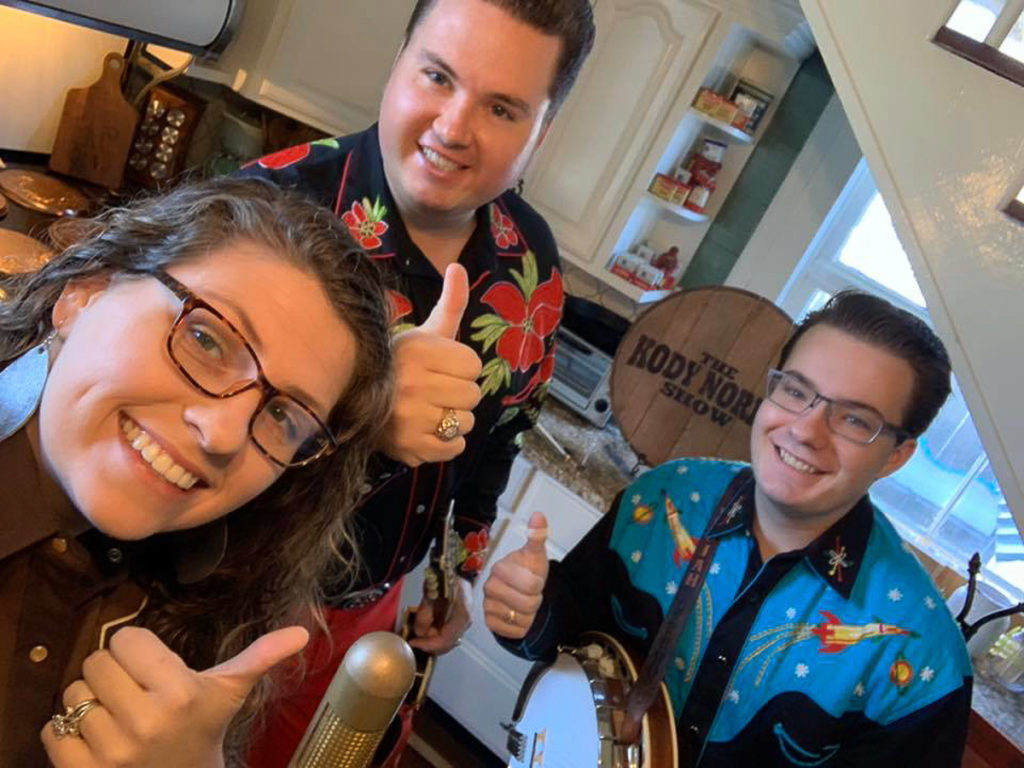 Three members of The Kody Norris Show around a mic in their kitchen, ready to record from home.