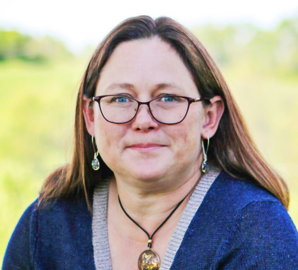 Author photograph: Headshot of KBB wearing a blue sweater, dangly earrings, a large necklace, and glasses.