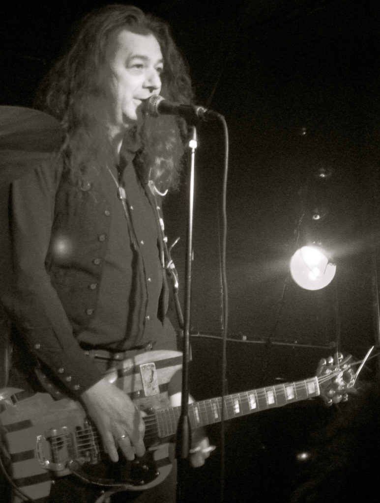 Fred Cole on stage singing at the mic and holding his guitar.