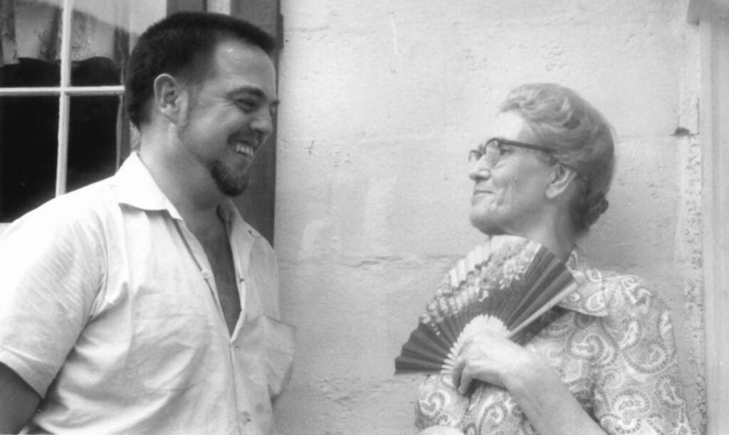 This black-and-white image shows a young man with dark hair and a short beard with an older woman with her grey hair pulled back in a bun at the nape of her neck. The man is smiling at the woman, and she looks at him with a small smile and holds a fan below her chin.