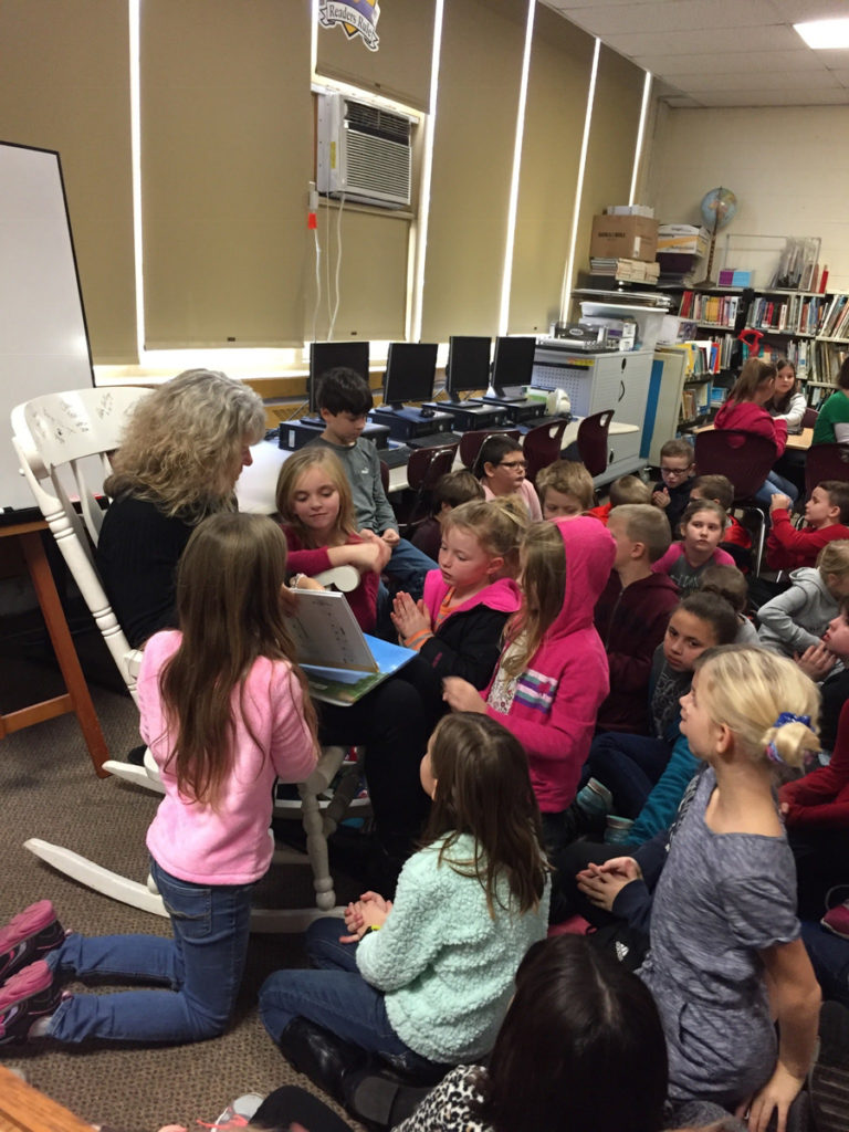 Nancy sitting in a rocking chair in a school classroom, surrounded by 2nd-grade students as she reads to them.