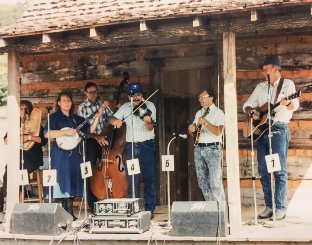 Several musicians on a log cabin front porch stage playing hammer dulcimer, banjo (Dee-Dee Price), bass, fiddle (Dean Sturgill), mandolin, and guitar.