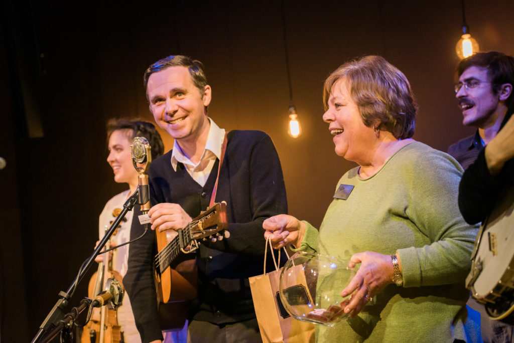 Kris Truelsen announces the winner of the maple syrup at the mic, with Leah Ross holding the jar of tickets and the gift bag waiting for the winner.