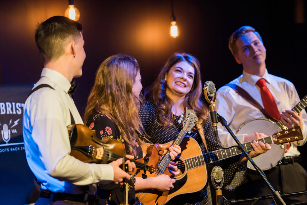 The four band members, with the male musicians on either side and the two female musicians in the center, gather round the mic with their instruments.
