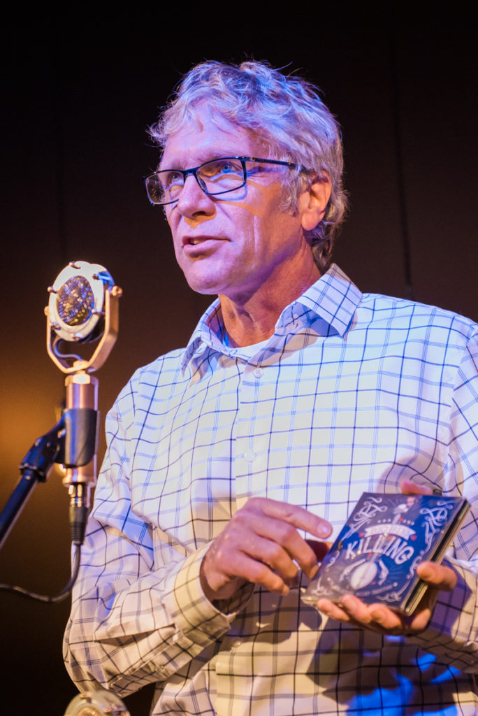 Ted standing in front of the microphone, holding the CD Big Bend Killing.