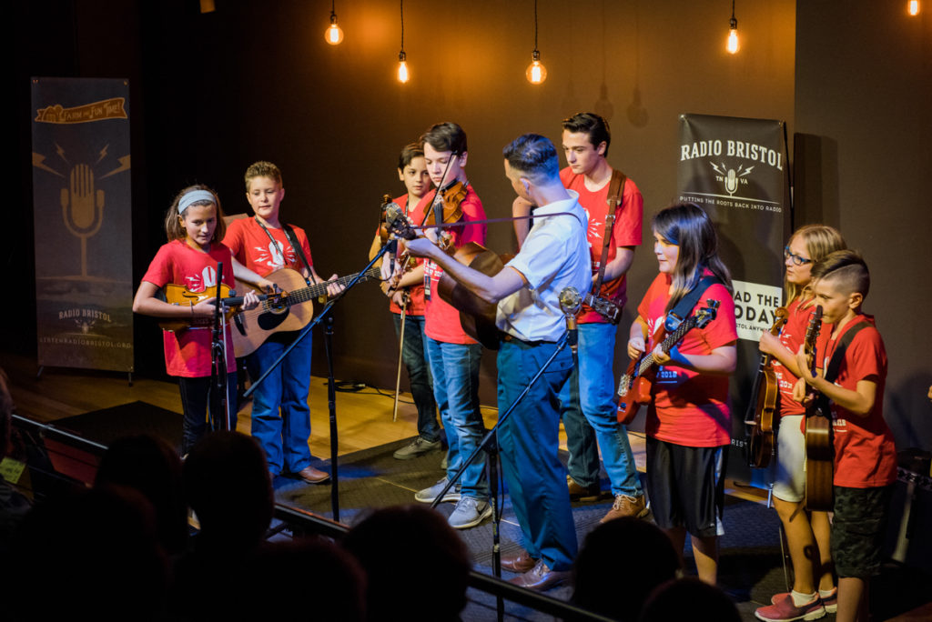 A shot of the Pick Along Summer campers on stage with Kris Truelsen; the kids are all wearing their red Pick Along logo shirts and have their instruments with them, ready to play.