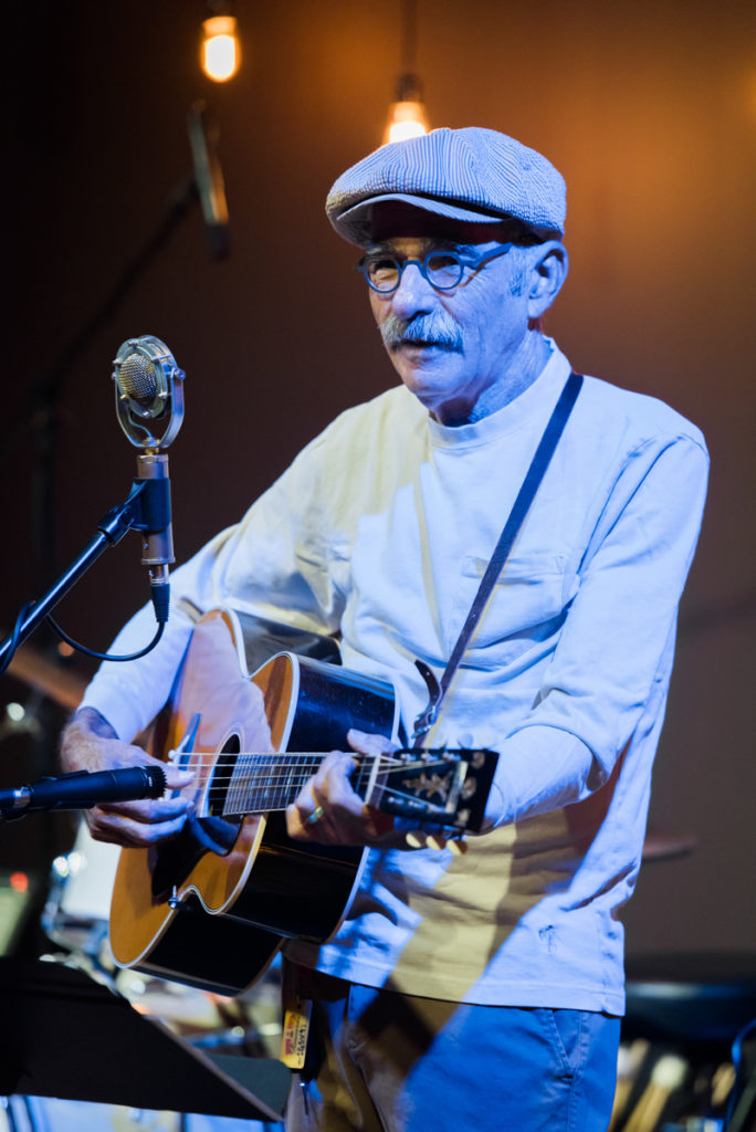 Roy Book Binder at the microphone playing his guitar and singing.