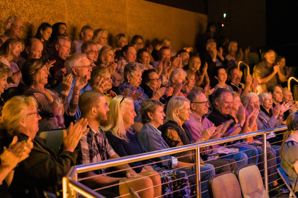 The crowded theater at October's show with row upon row of audience members clapping.