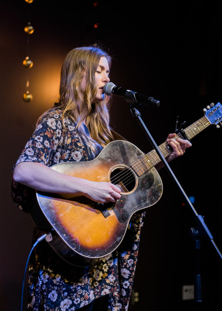 Jill Andrews holding guitar and singing at microphone