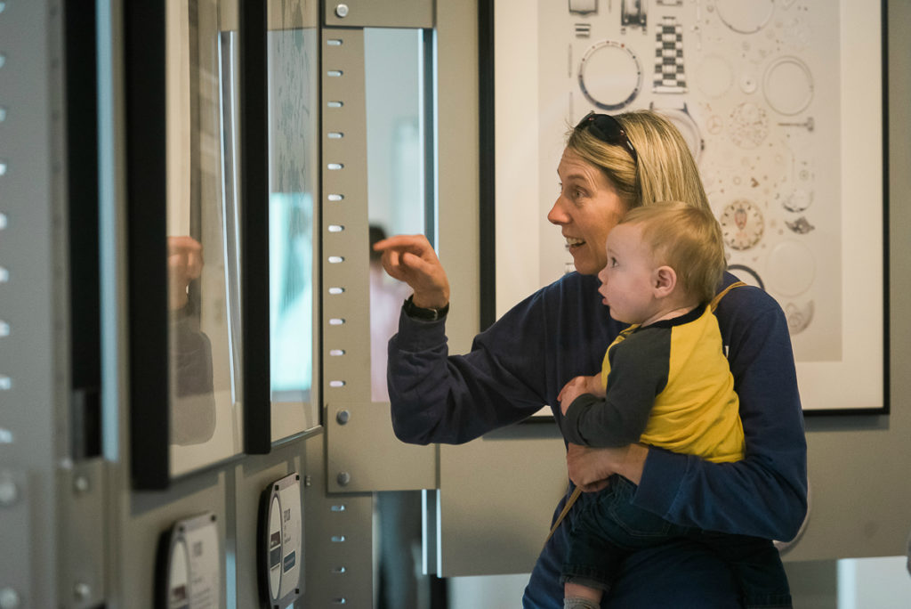 Photo of blond woman holding a baby and pointing at an image on the museum's wall. Both of them have expressions of wonder on their faces.