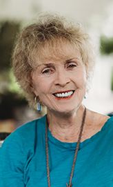 Photograph of a woman with short blonde hair, smilling and wearing a turquoise shirt and earrings.
