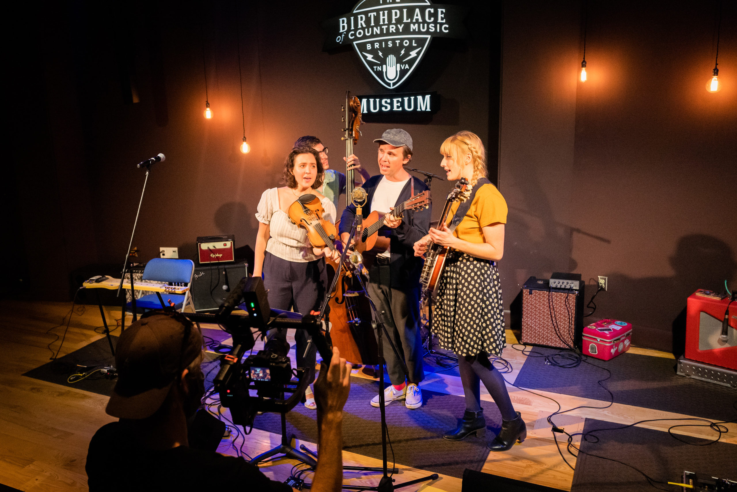 Three musicians -- female fiddler, male guitarist, female banjo player -- playing and singing at a mic with the male bass player behind them. There is a man with a large video camera filming them in the foreground.