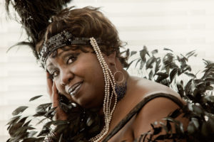 A Black woman looking sideways at the camera, seen from the chest up. She is wearing a black thin-strapped dress with a black and pearly headdress with feathers and other adornments. She has short hair.