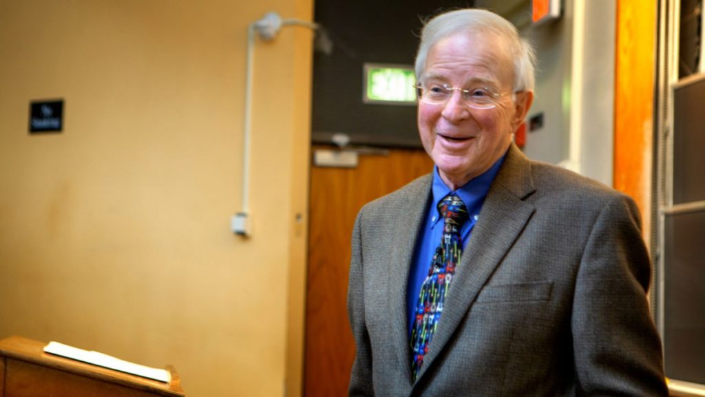 In this image, an older white man with white hair stands in a lecture theater near a podium. He is wearing a grey suit jacket, a royal blue shirt, and a colorful tie; he also wears glasses.
