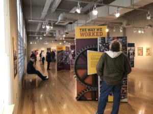 A view of the Special Exhibits Gallery with visitors exploring the stand-up panel modules for The Way We Worked. In the foreground, a man with a dark jacket and a hoodie stands in front of the opening panel.