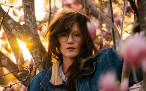 A photo of S.G. Goodman standing in front of fall trees with the sunset peaking out just over her shoulder.