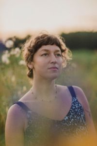 Photograph of a young white woman with brown curly hair pulled back at the nape of her neck. She is wearing a sleeveless blue-patterned top and a delicate beaded necklace. She is in a field with flowers behind her.