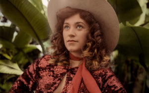 A photo of Sierra Ferrell set against a backdrop of foliage. She is wearing a cowboy hat and vintage western-wear.