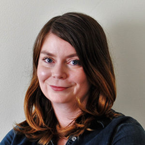 This photograph is a headshot of a white woman. She has reddish brown hair that reaches to her shoulders, and she is wearing a blue button-down shirt. Her eyes look blue and she is smilling at the camera.