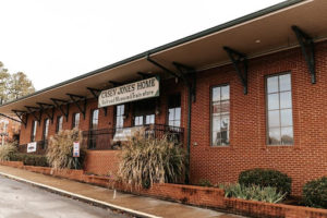 A long single-story brick building with garden planting in front of it and the museum's name on a sign before the entrance door.