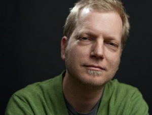 Color image of a white man shown from the upper chest/shoulders up. He has short light brown/dark blonde hair and a "soul patch" tuft of hair below his lower lip. He is wearing a green shirt.