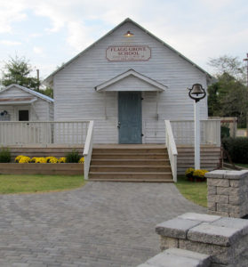 A white clapboard building -- an old schoolhouse -- with steps leading up to a pale blue grey door. There is a bell on a pole near the steps.