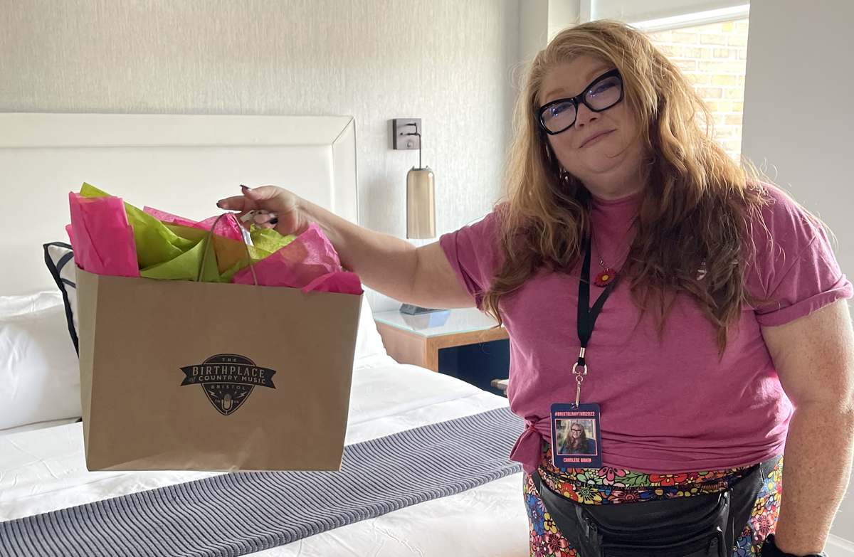 Photo of Charlene Baker standing in Rosanne Cash's suite at The Bristol Hotel, holding a gift bag.