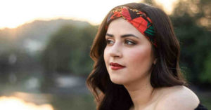 A close-up of Florencia from the shoulders up -- she has long brown hair and is wearing a red patterned hair band/scarf to hold her hair back from her face. The background is blurred but seems to be trees and buildings. 