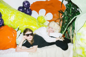 Image: Two white women lie in the center of the image looking at the camera. The woman in the foreground is wearing all black and sunglasses; she has medium-length brown hair. The woman behind her is wearing all white and has platinum blond hair. Vibrantly colored fruits and flowers are painted on the background behind them.