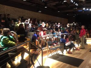 A large theater space with school students seated while a white man in a red shirt and khaki pants talks to them from the stage floor.