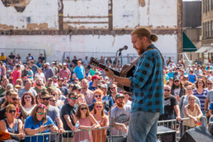 Tyler Childers is on stage and faces a crowd of fans watching him as he performs. He is wearing a black and blue plaid shirt playing a guitar looking down and singing. It is a bright and sunny day.