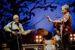 97 year old Clyde Lloyd looks onward toward Tyler Childers as they play onstage. Both are playing guitars in front of a stage backlit by a blue backdrop with a large silhouette of a tree behind them.