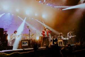 Musician Tyler Childers singing into a microphone with a full band, a bass player to his right, a pedal steel guitar and electric guitar players to his right. Bright stage lights are shining down upon the band as they play and sign passionately.