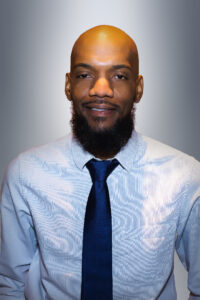 A Black man in a light-colored shirt and dark tie. He is bald with a trimmed beard, and he is looking directly at the camera.