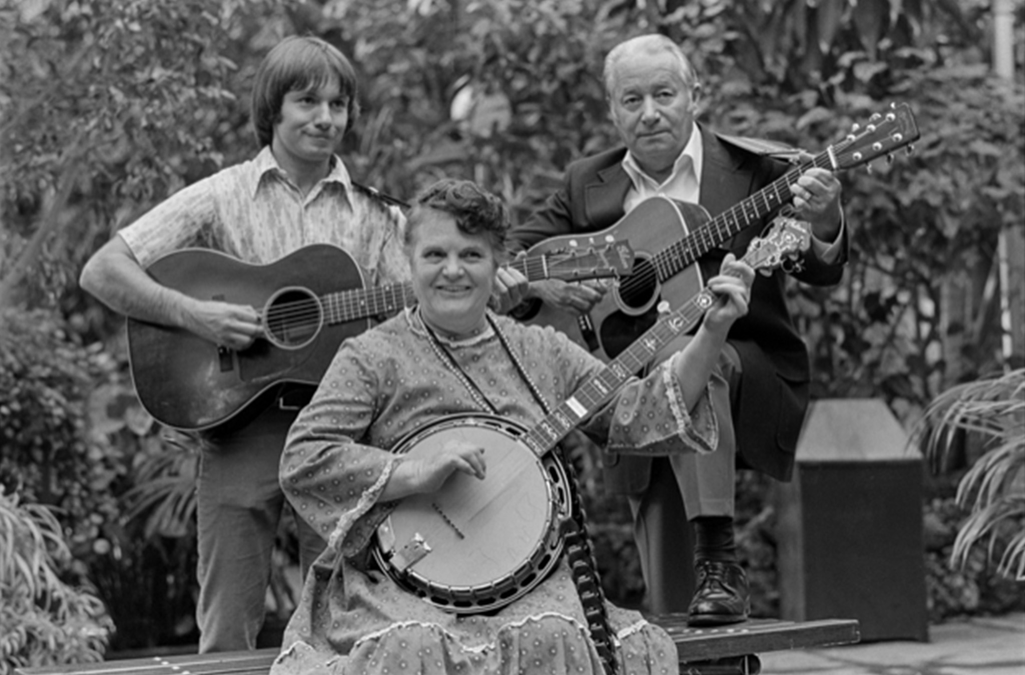 Black-and-white photograph of three musicians: a young man with light brown hair, standing with guitar; a middle-aged woman in old-fashioned clothes, sitting with banjo (Ola Belle Reed); and an older man in a dark jacket standing with a guitar.