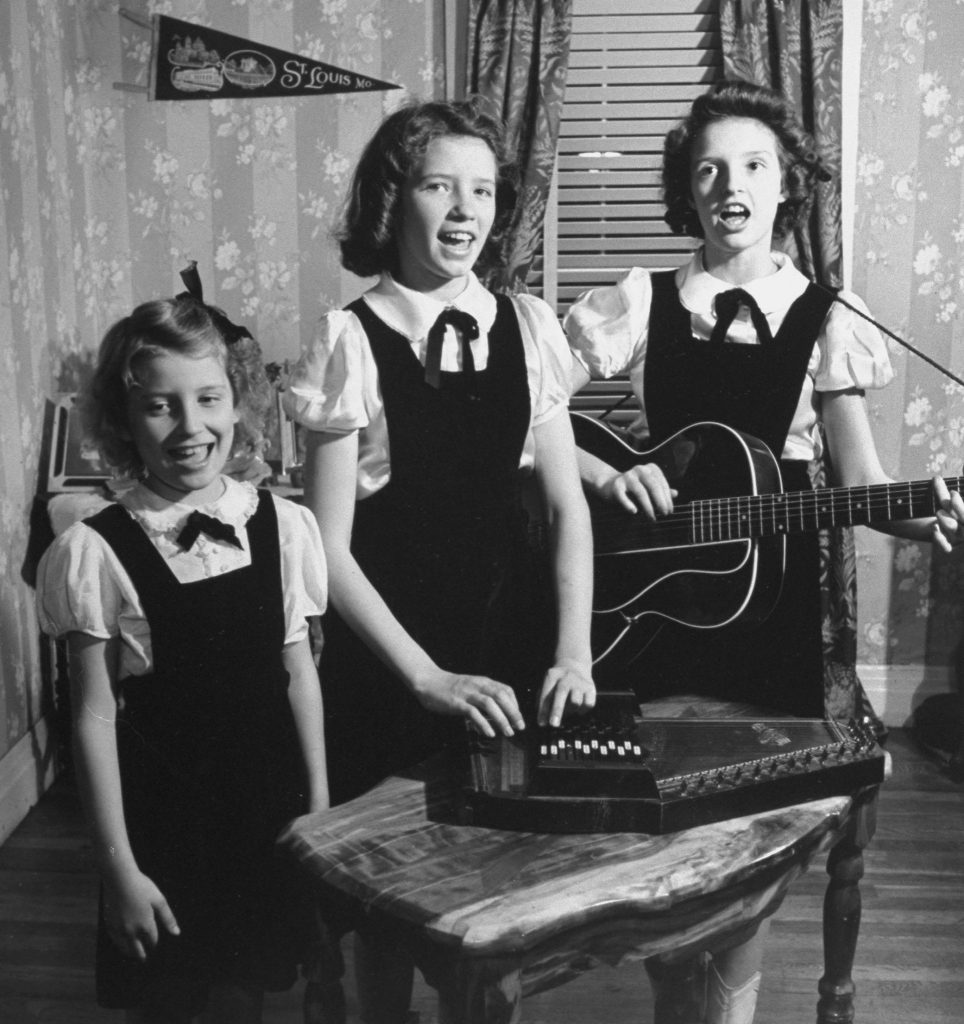 A black and white image of three young girls, Anita, June and Helen Carter. The Girls are singing and dressed in the same overall dress outfit with bows at their necks. Helen is standing, singing and playing a guitar, June is in the middle touching an autoharp on a table in front of her, and Anita is smiling and singing.