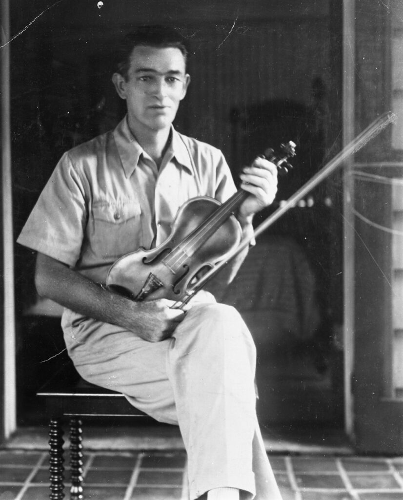 A black and white image of Charlie Bowman. He is seated on a small bench and holding a fiddle in his lap. He is wearing a collared shirt. The image is old and not completely clear, his face is slightly fuzzy.