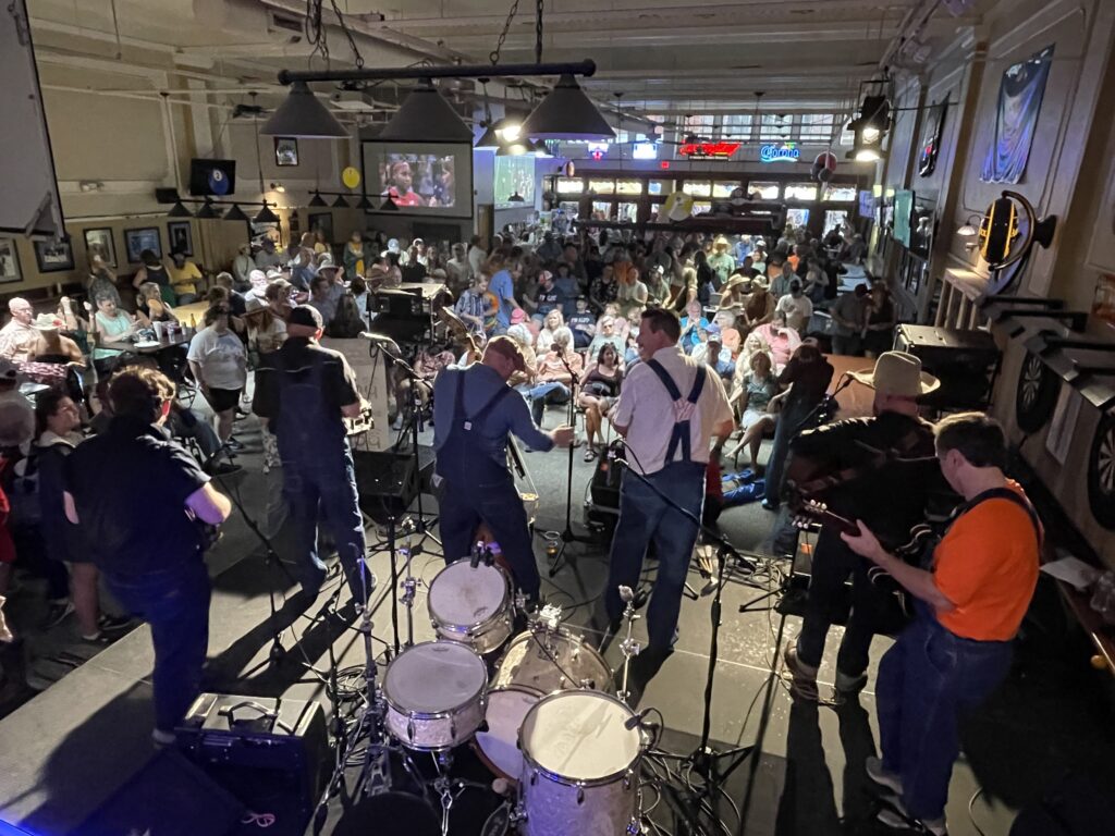 Photo taken from behind the band Old Line Skiffle Combo as they perform on the Borderline Billiards Stage at Bristol Rhythm & Roots Reunion. Before them is a large audience, filling the room.