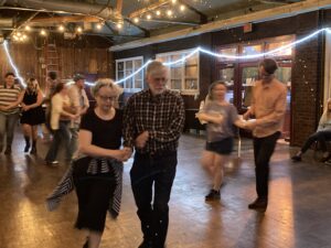 Several paired square dancers move in a large room with fairy lights and other fun decorations above them.
