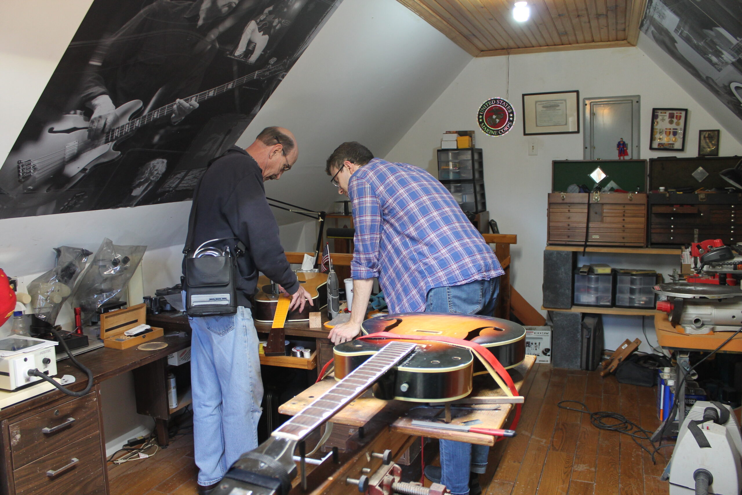 Two men with their backs facing the camera inspect a broken guitar inside of a small guitar repair shop. The room is filled with tools and instruments.