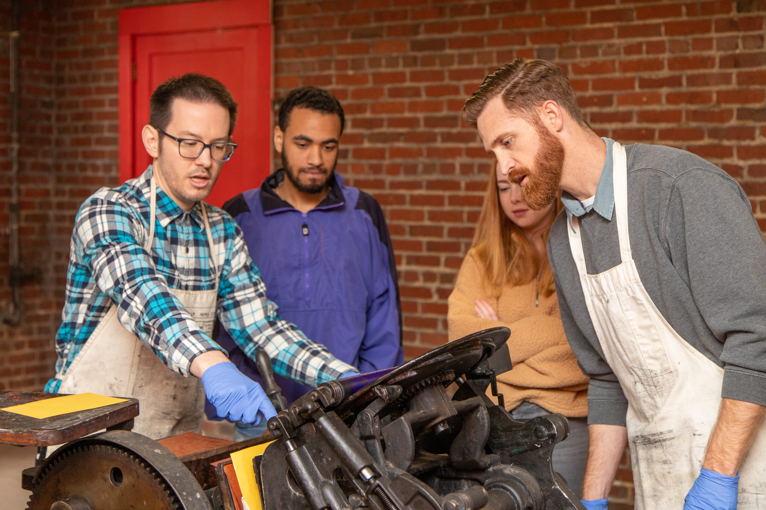 Four people are standing around a vintage printing press, looking at the press as one man operates it. 