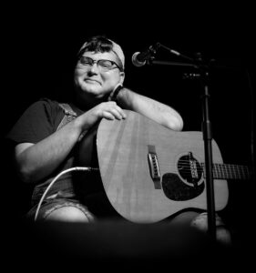 White man with backwards ballcap, glasses, and overalls sitting in front of a mic stand. A guitar sits on his lap and he is smiling and resting his head on one arm propped on the guitar. 