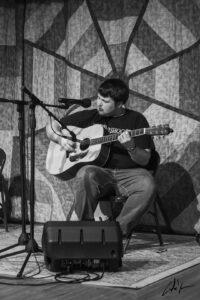 A white man sits playing a guitar in front of a mic on a stage with a quilted backdrop.