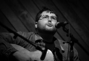 A white man with glasses and a jean jacket is singing into a mic and holding a guitar.