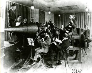 An image of men playing in an acoustic recording session. Multiple musicians sit crowded around a large metal horn, playing directly into it so it captures their sound.